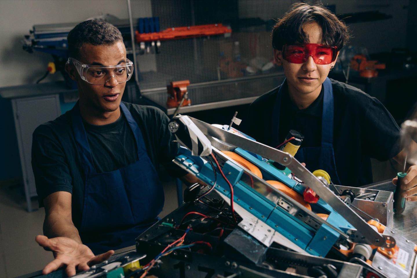 Two device engineers in safety glasses working through a partially disassembled electronic device