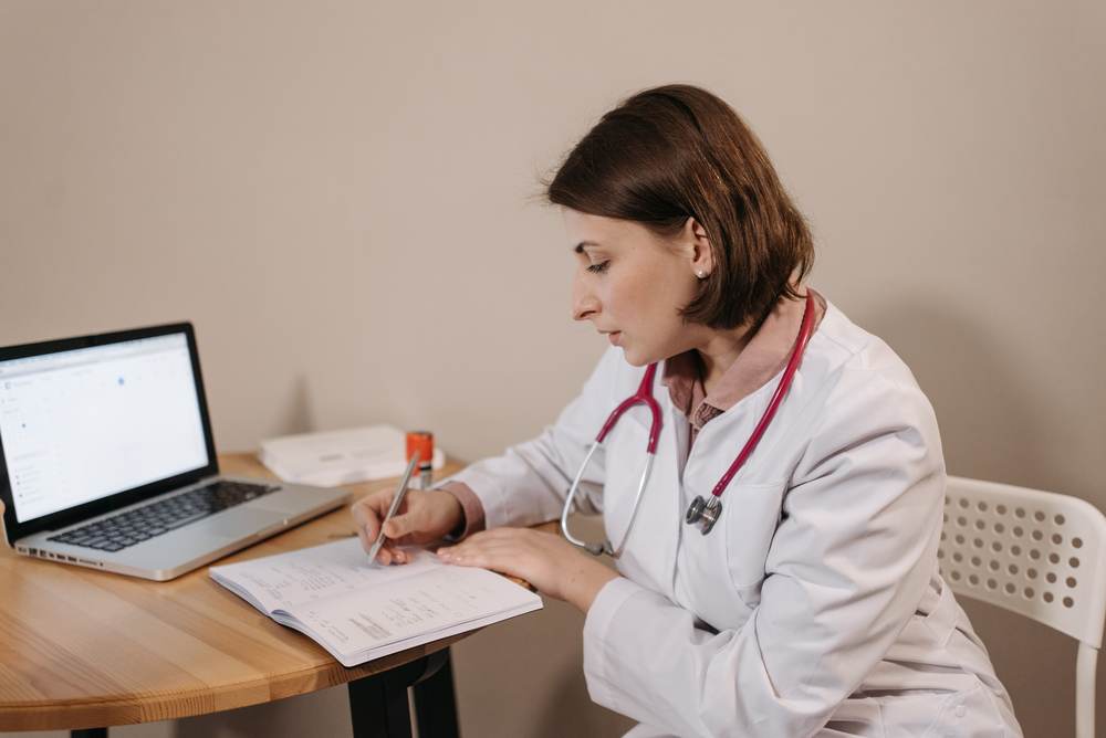 A doctor in a clinic office writing a prescription beside a laptop