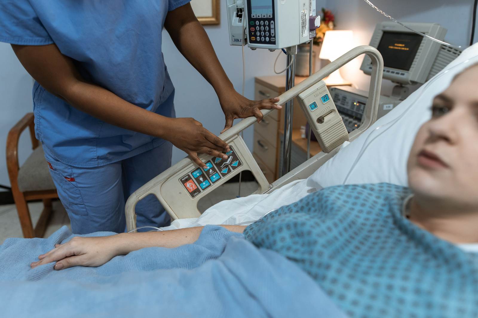 A nurse in scrubs assists a patient in a hospital bed with medical equipment nearby