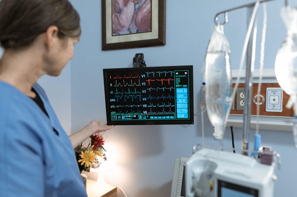 A nurse in blue scrubs examines a medical monitor displaying vital signs in a hospital setting
