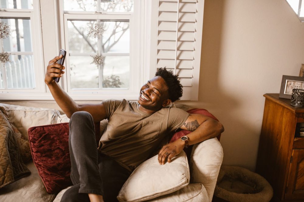 A smiling man on a sofa taking a video call on his phone at home