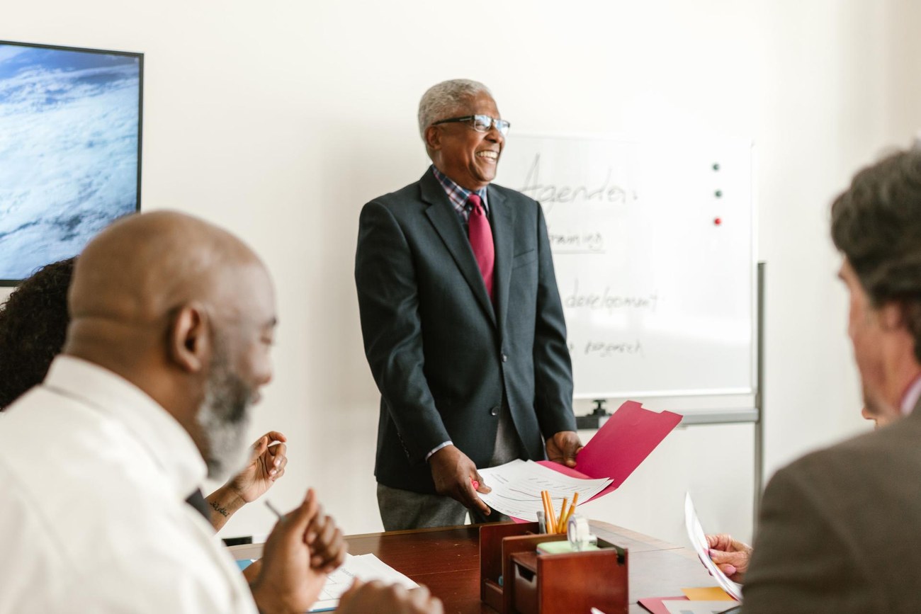 Senior presenter smiling while handing a folder of papers to colleagues at a meeting table, whiteboard visible behind him