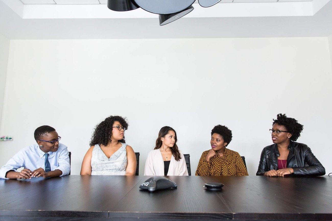 Five colleagues seated around a conference table in a bright white-walled meeting room, in conversation