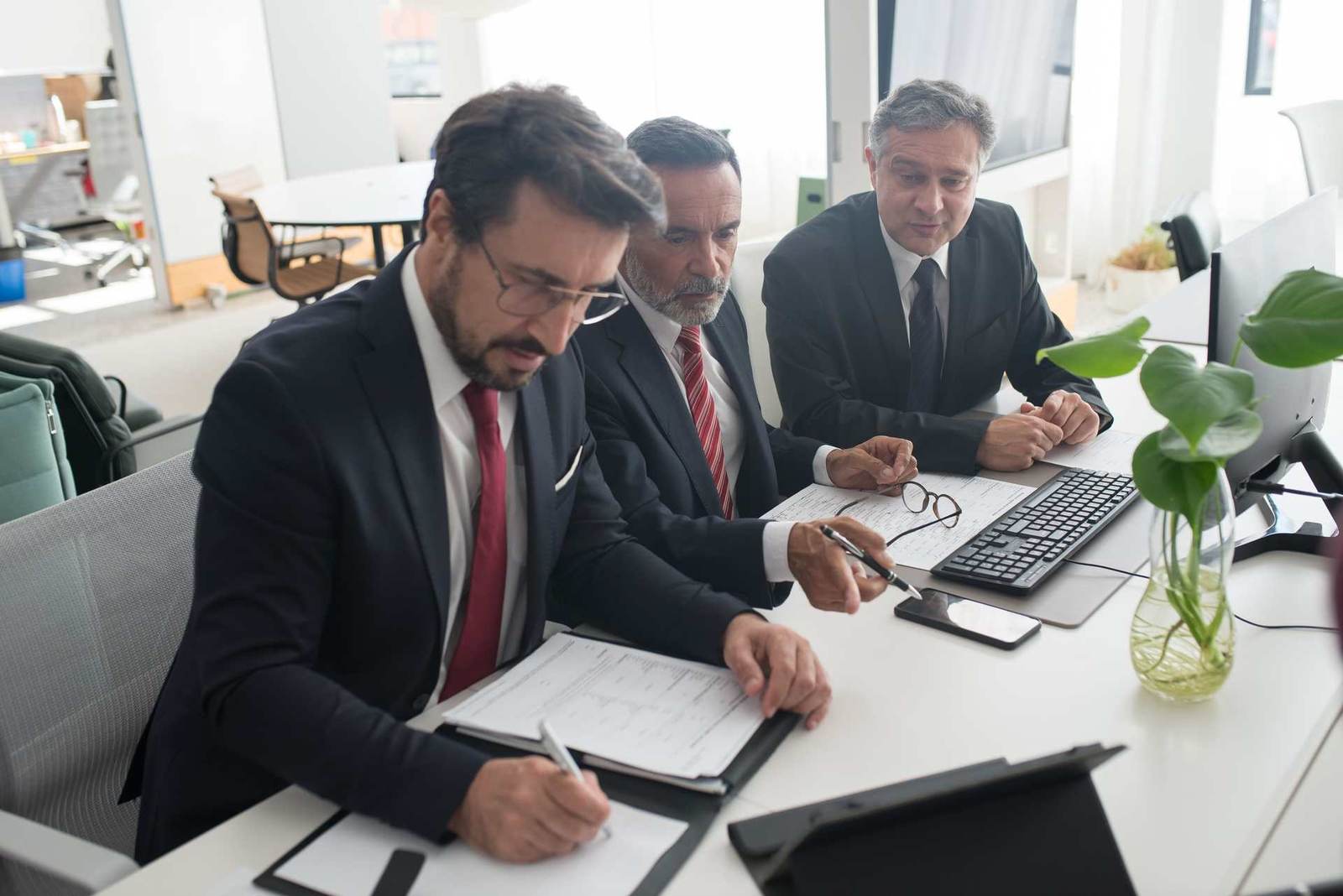 Three colleagues in suits review a marked-up technical document together at a desk with a keyboard and monitor