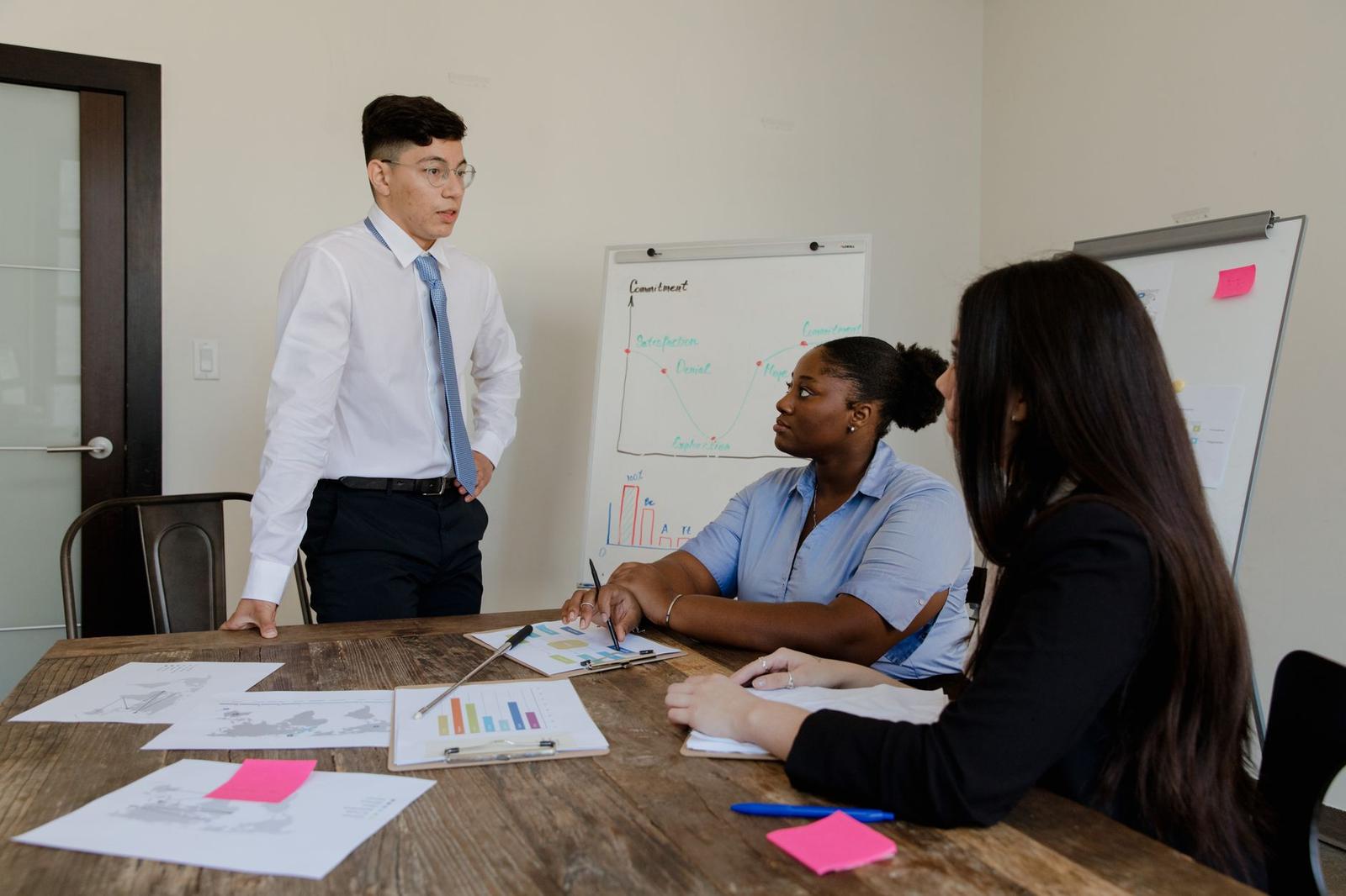 Colleagues gathered around a conference table reviewing charts and diagrams during a strategy discussion, one standing at a whiteboard