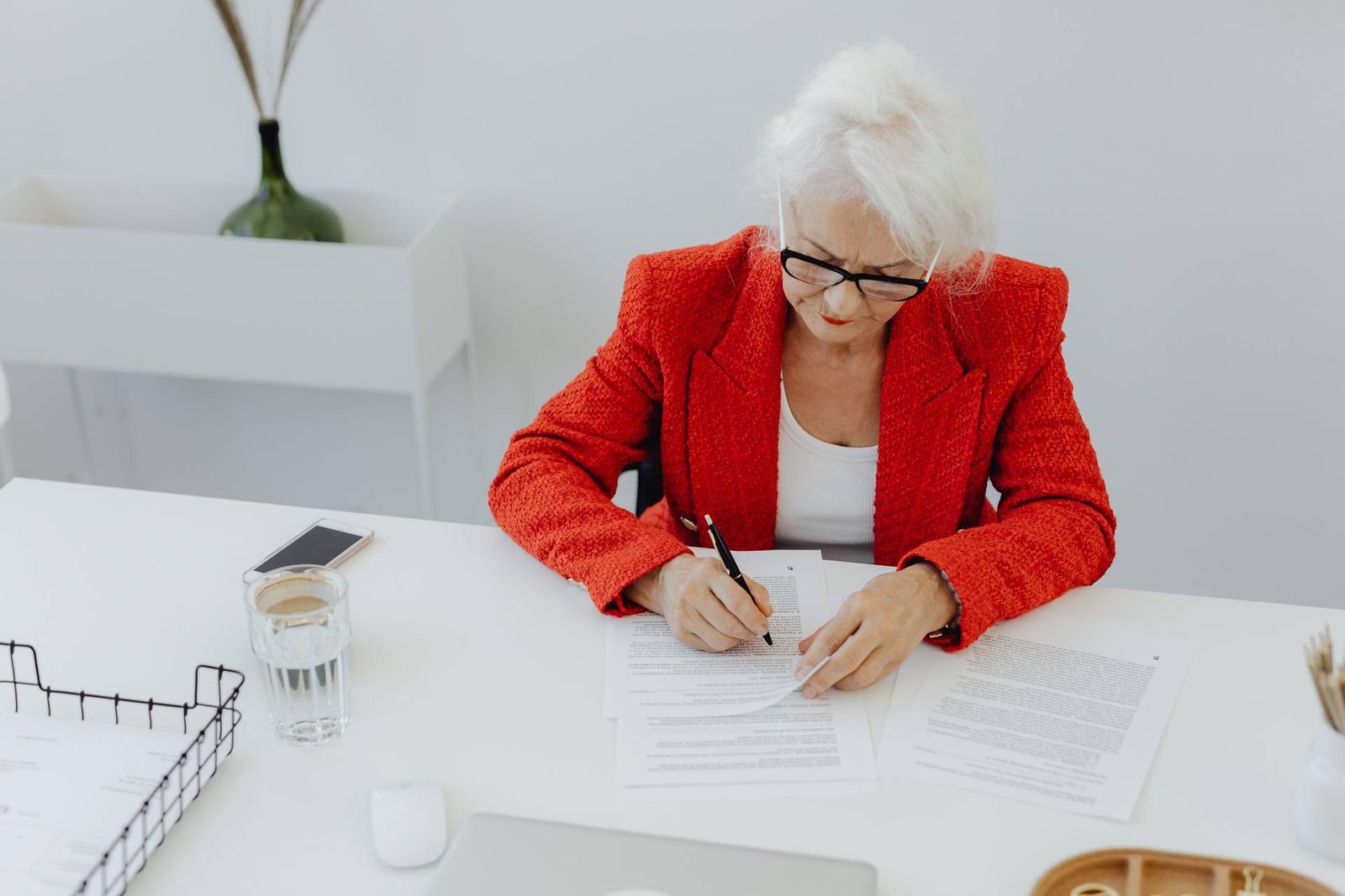 Senior professional in a red blazer signing a document at a clean white desk