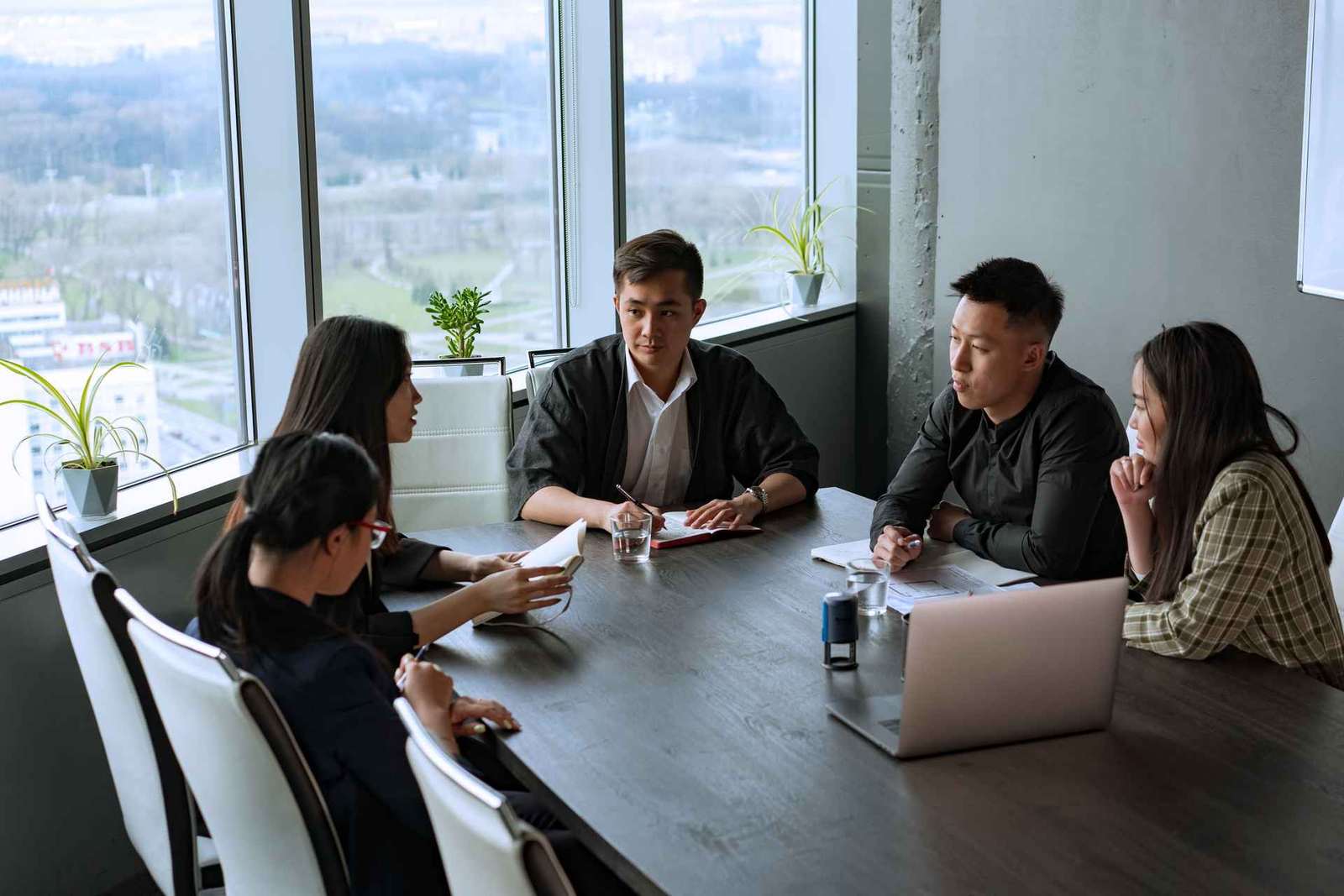 Five colleagues gathered around a boardroom table with a laptop, in discussion with a city and park view through floor-to-ceiling windows