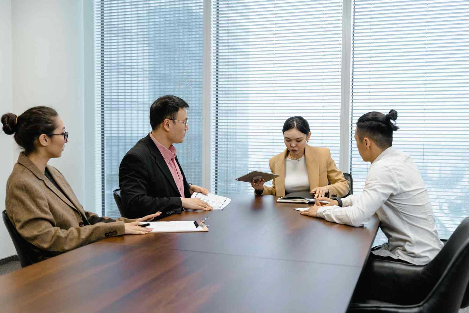 Four colleagues seated around a boardroom table reviewing documents and a tablet together in a bright, window-lit conference room