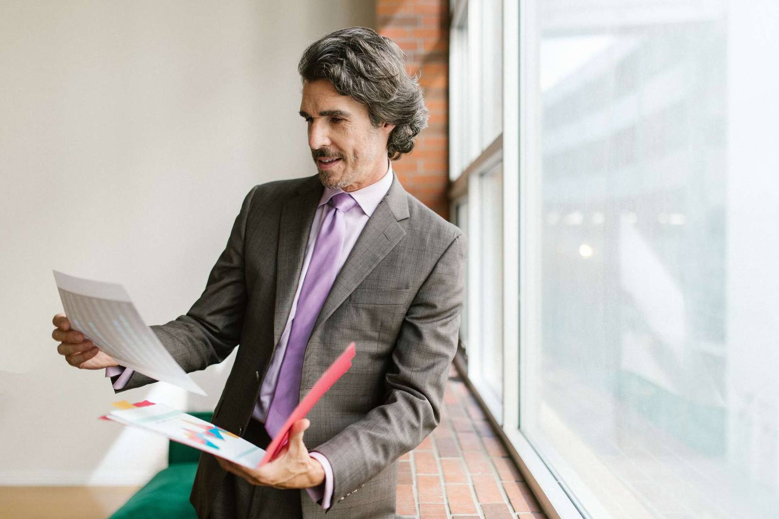 A man in a suit reviews a printed report and a color-coded folder beside a tall office window