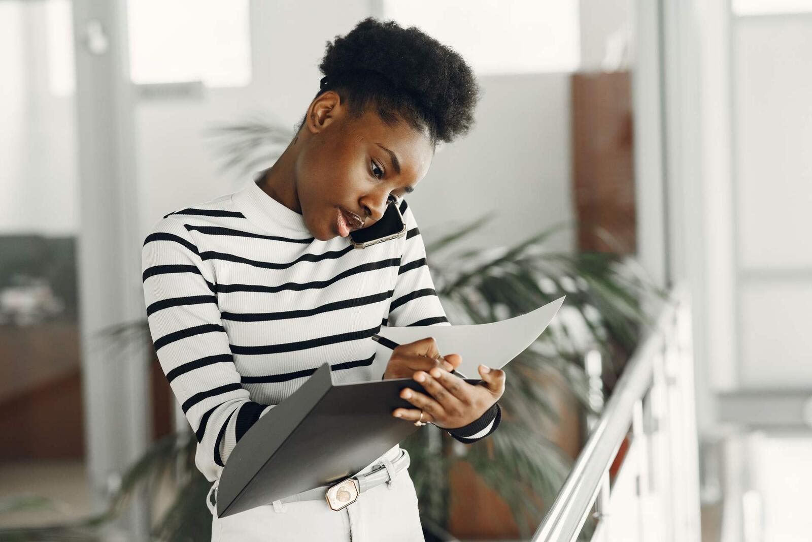 A professional woman takes notes on a clipboard while on a phone call in a bright, plant-filled office atrium
