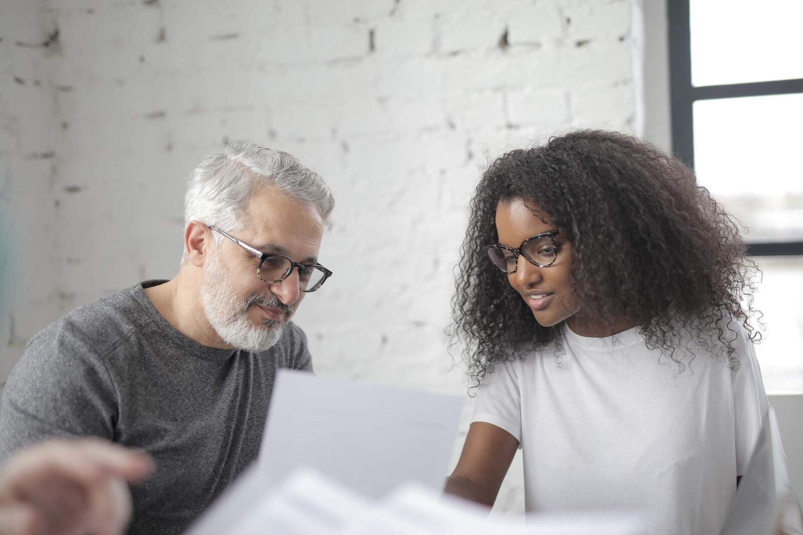 Two colleagues reviewing a printed document together against a white brick wall