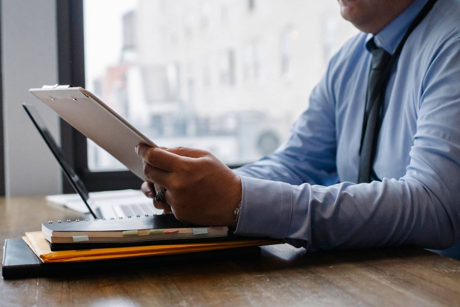 Professional reviewing a clipboard document beside an open laptop, city buildings visible through the window