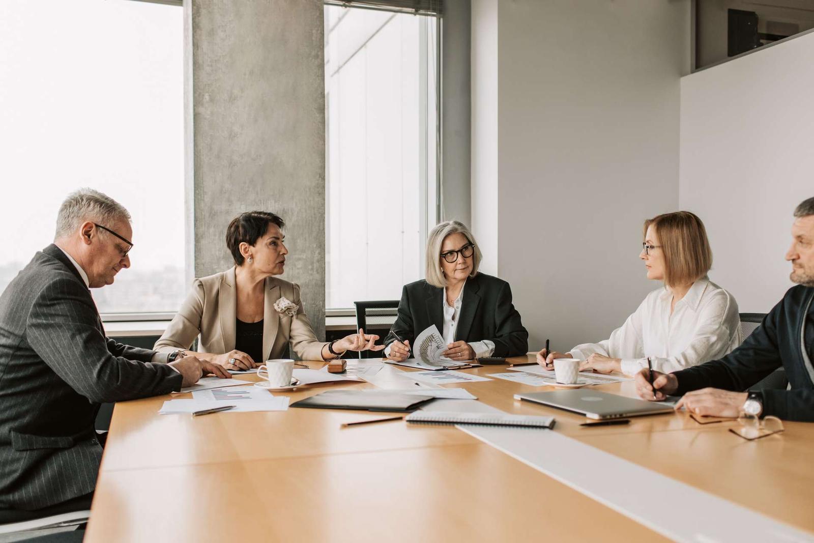 Five senior colleagues review printed charts together around a bright conference table by a window