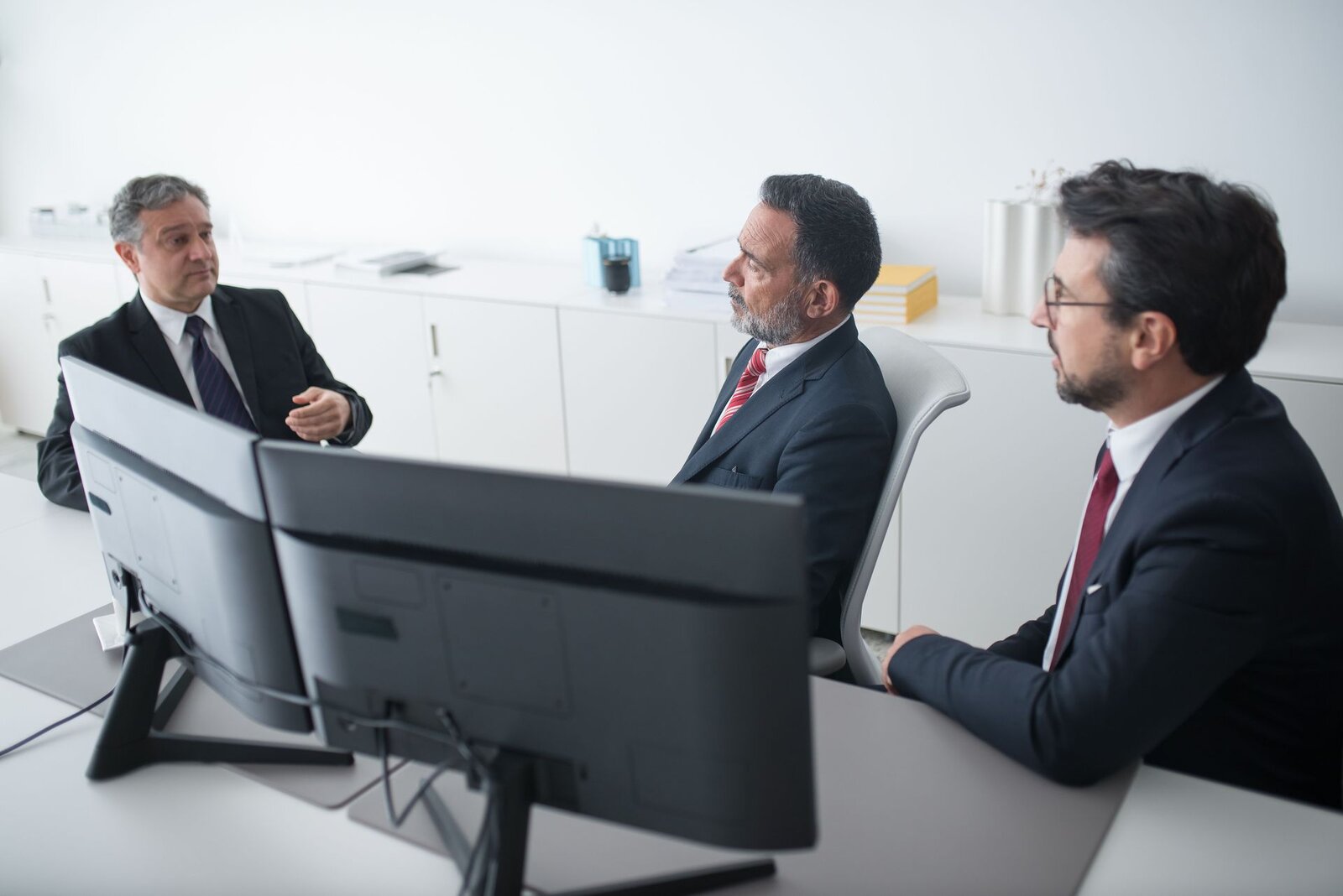Three regulatory colleagues in dark suits discussing documents at a desk in a bright white office