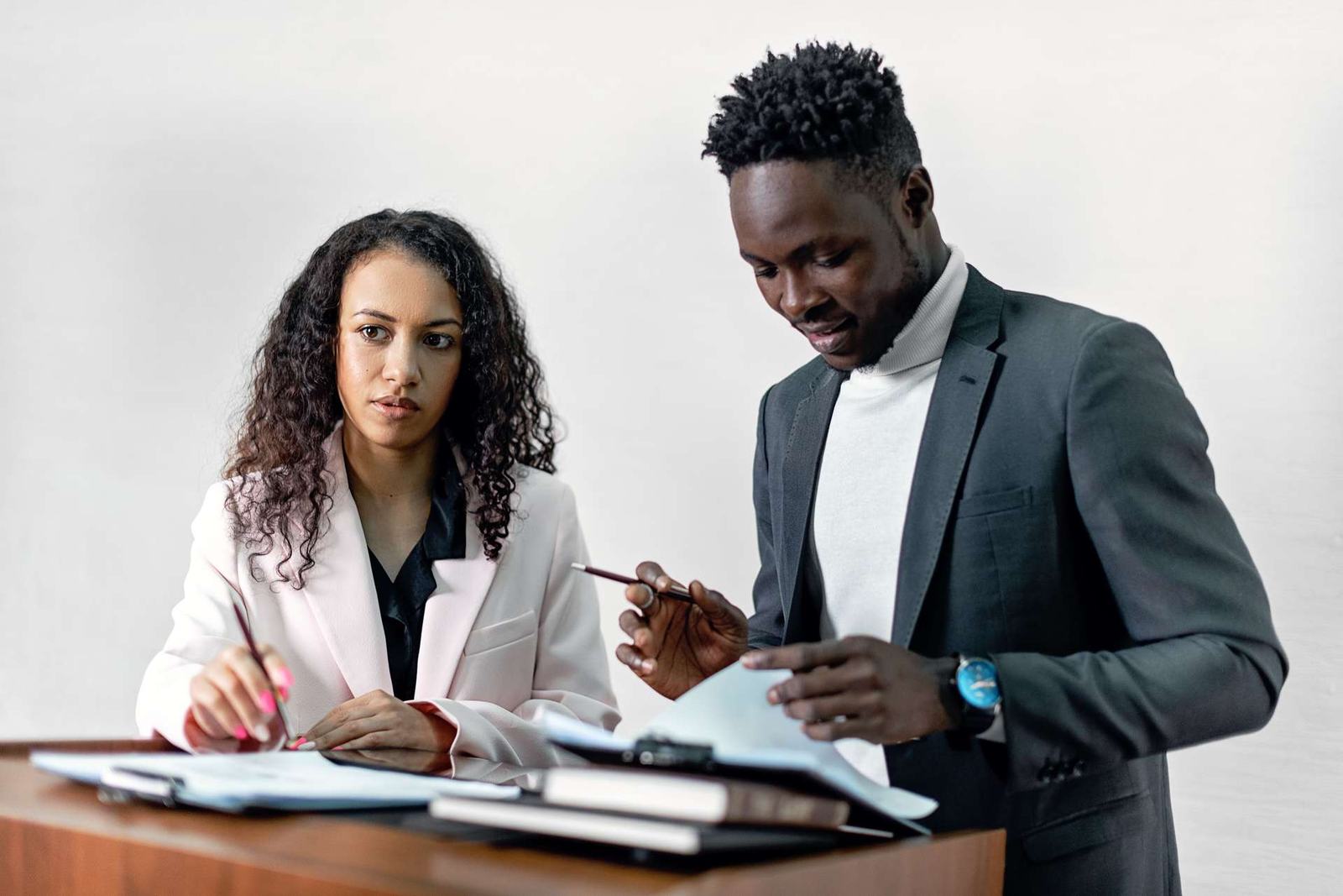 A woman in a blush blazer and a man in a dark suit reviewing printed reports together at a wooden desk