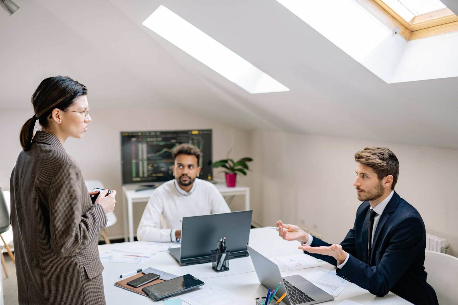 Three colleagues in a bright skylit office discussing data on laptops around a table