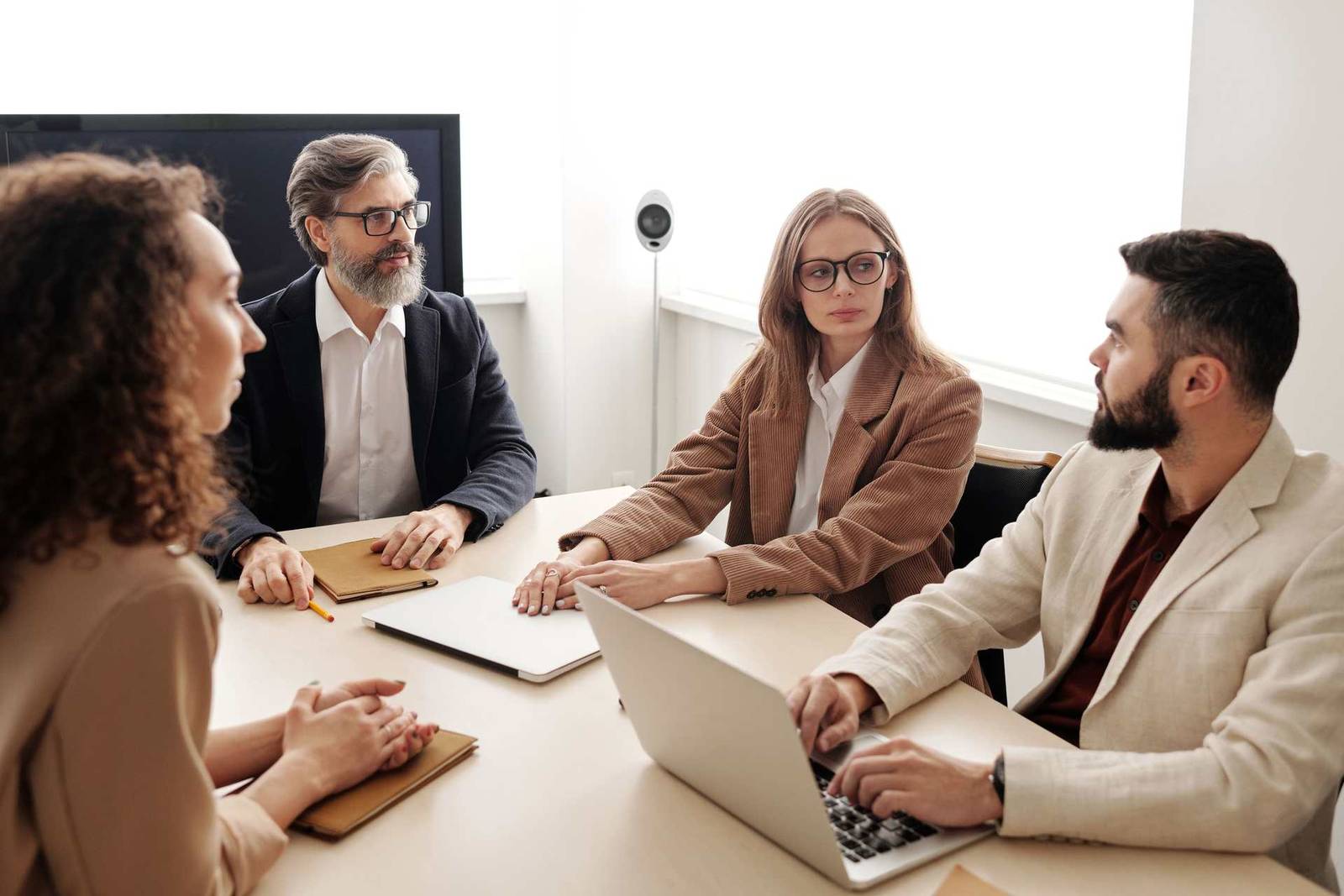 Four colleagues in a bright conference room, two reviewing a laptop while two others confer across the table