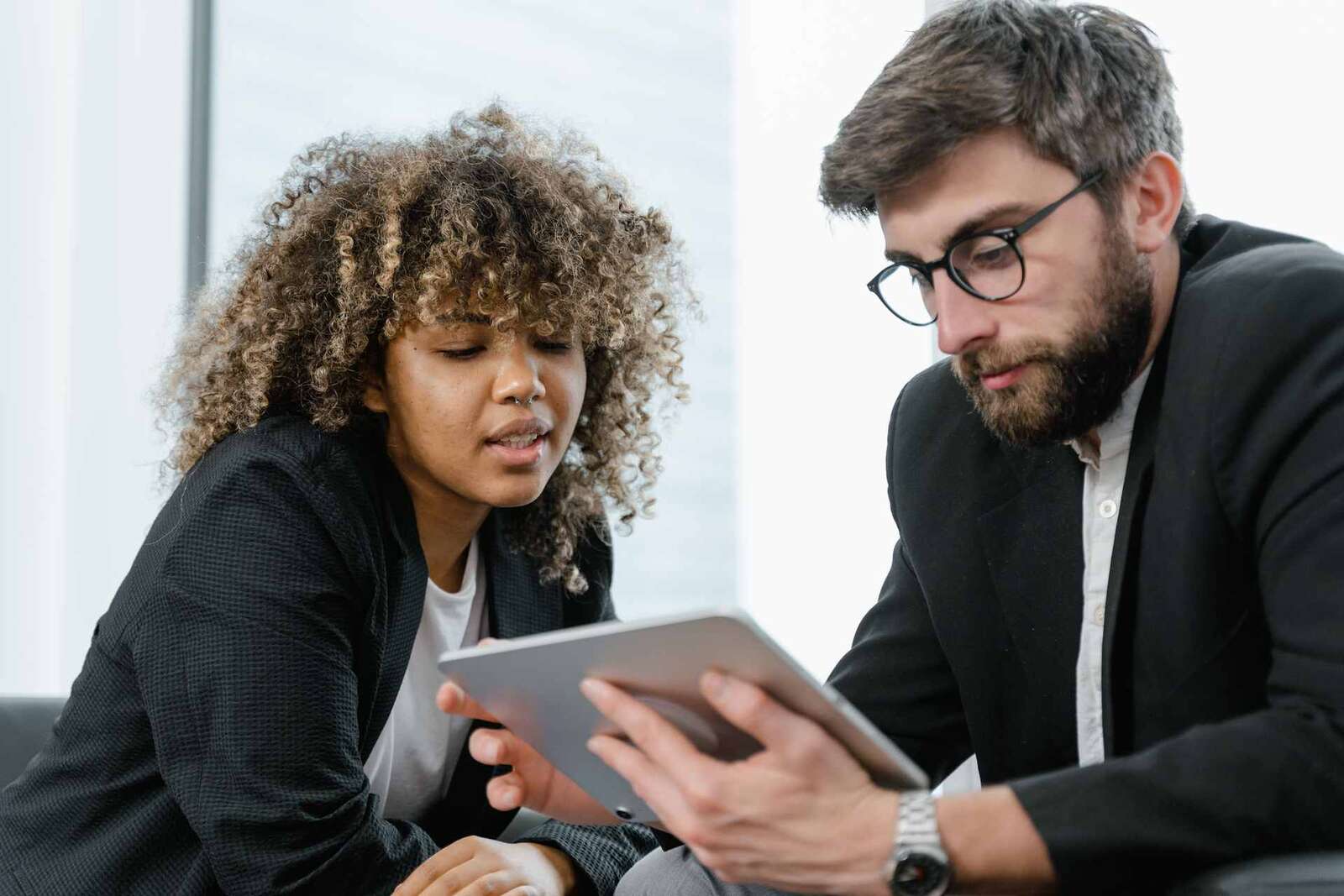 Two colleagues in blazers reviewing a tablet together by a bright office window