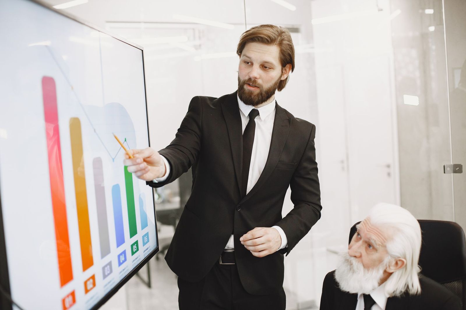 Man in a dark suit pointing at a colorful bar chart on a wall-mounted screen while an older, white-bearded colleague looks on in a bright glass-walled office