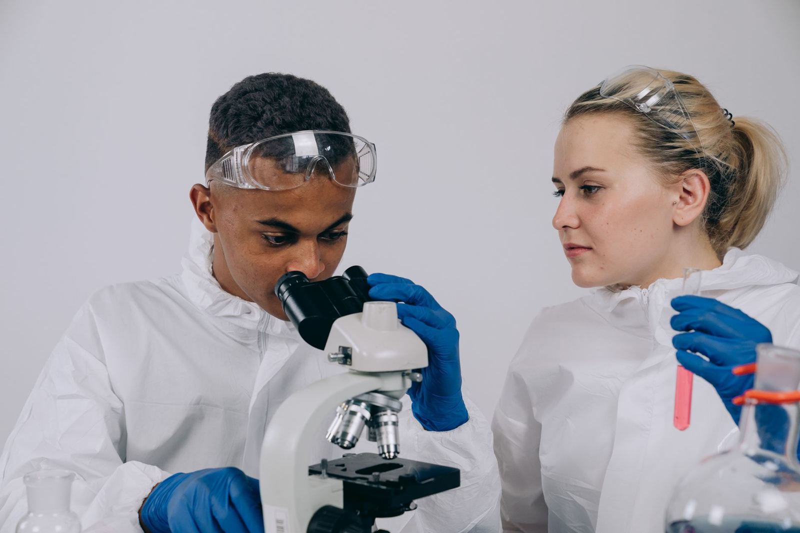 Two young scientists in lab coats and safety goggles examining a sample under a microscope