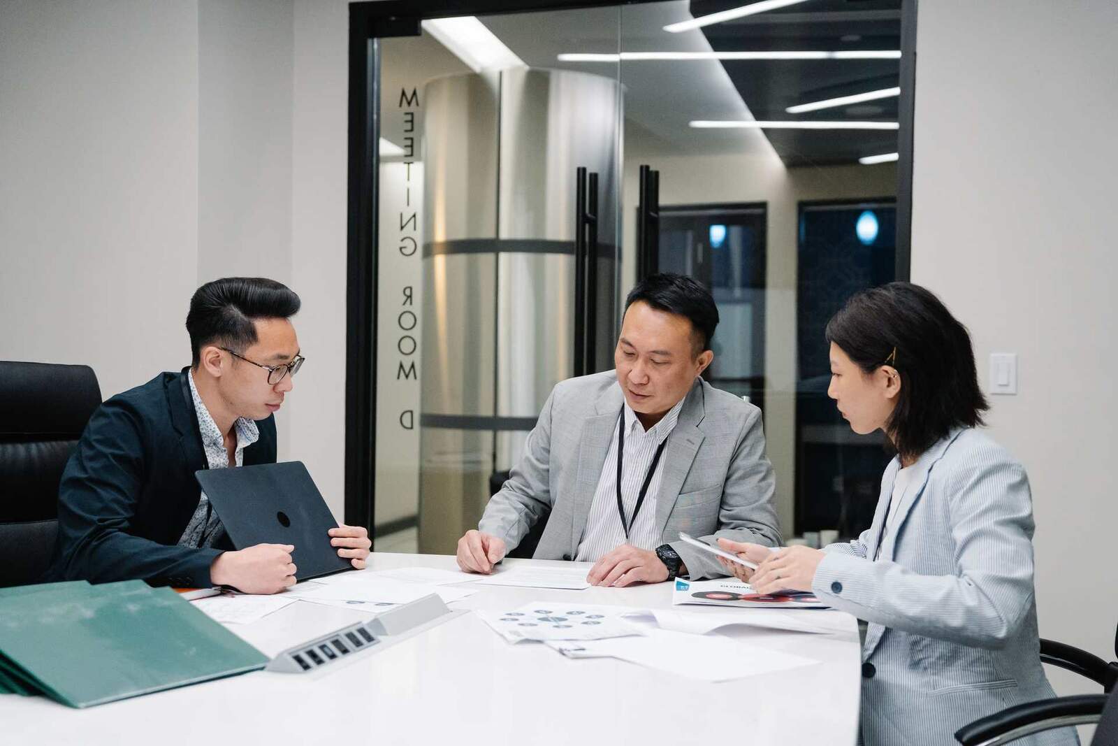 Three colleagues in dark blazers reviewing documents together around a table in a bright meeting room