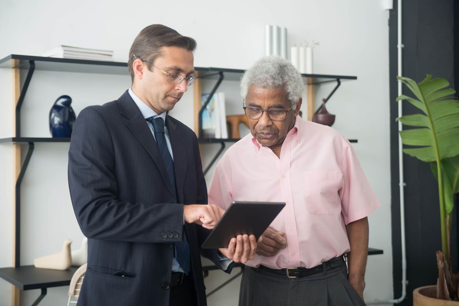 A regulatory advisor in a suit pointing to a tablet while explaining a document to an older client in a bright office with a bookshelf and a potted plant