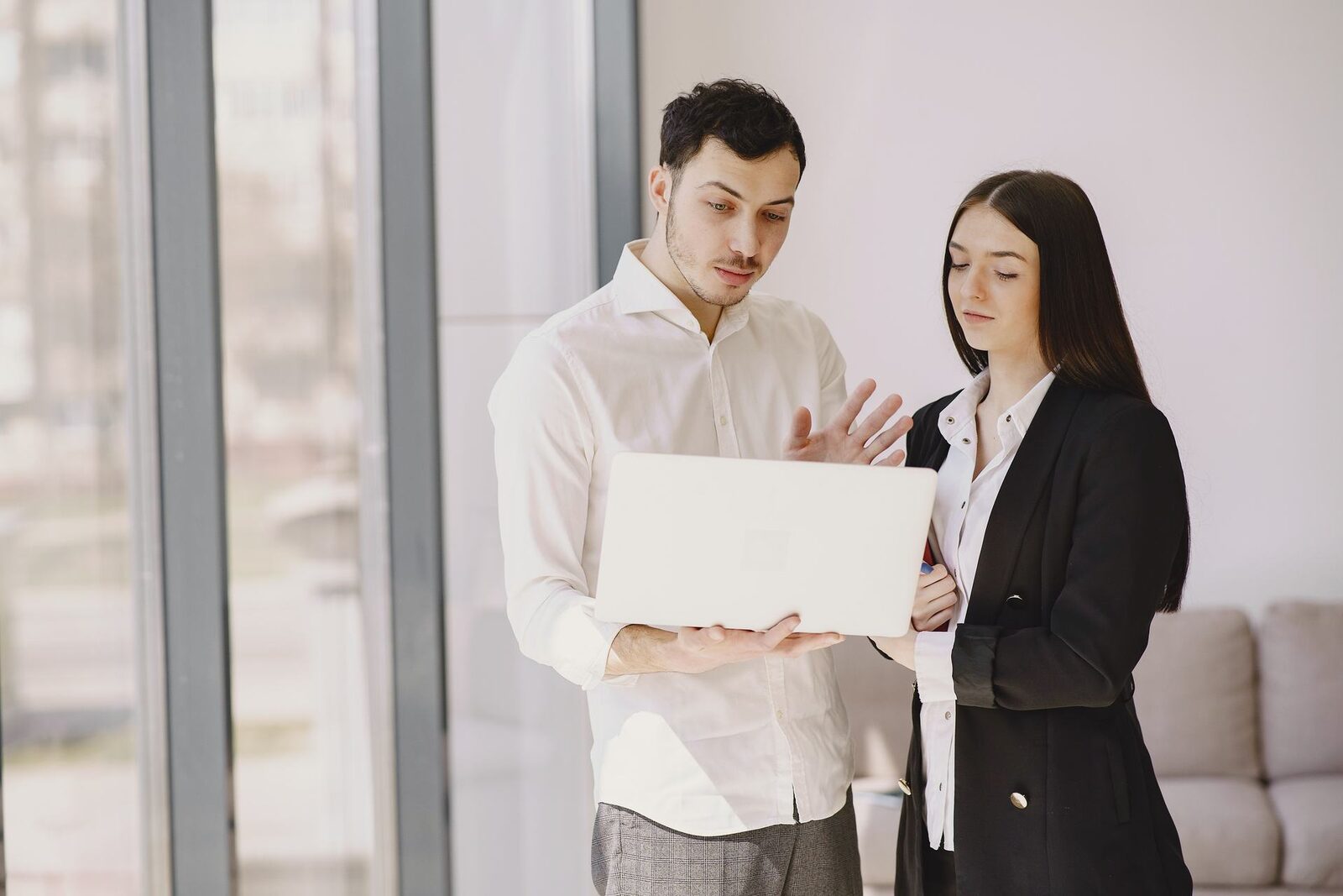 Two colleagues in business attire reviewing a document on a laptop in a bright office with floor-to-ceiling windows