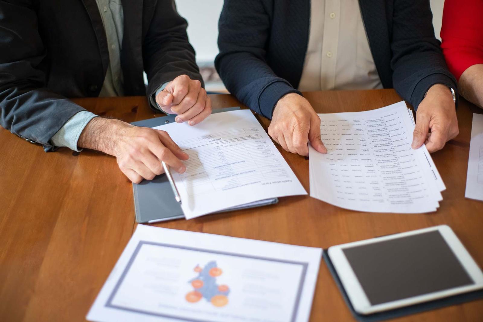 Two people's hands reviewing and annotating multi-page application forms on a wooden table