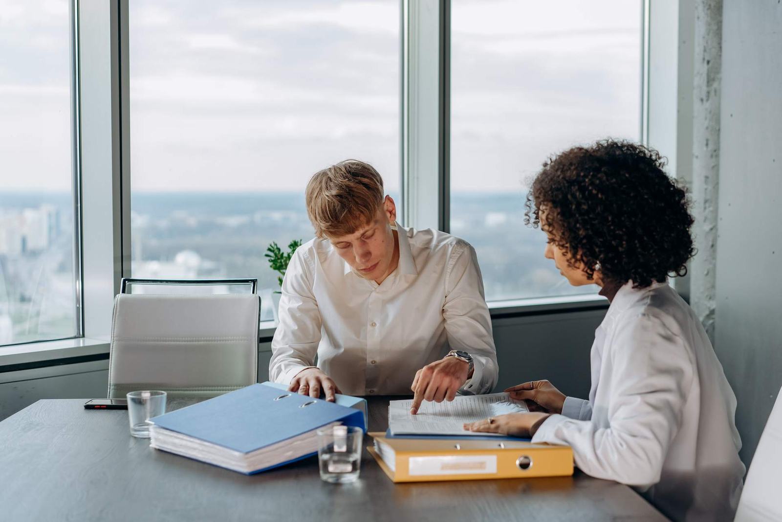 Two colleagues reviewing binders of documentation together at a high-rise conference table with a city view through floor-to-ceiling windows
