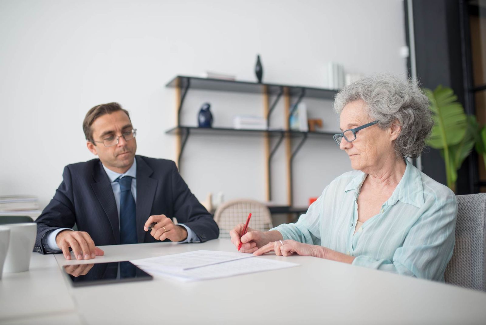A regulatory adviser and an older client reviewing and signing a document together at a desk in a bright office