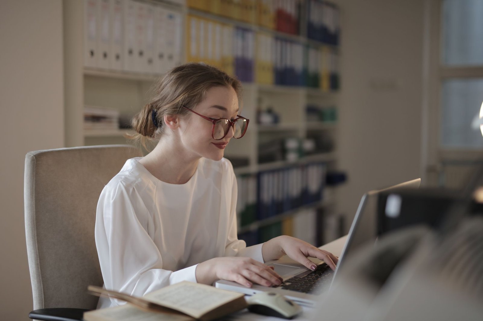 A regulatory professional reviews documentation on a laptop at a desk in a bright office, with binders of records on the shelving behind her.