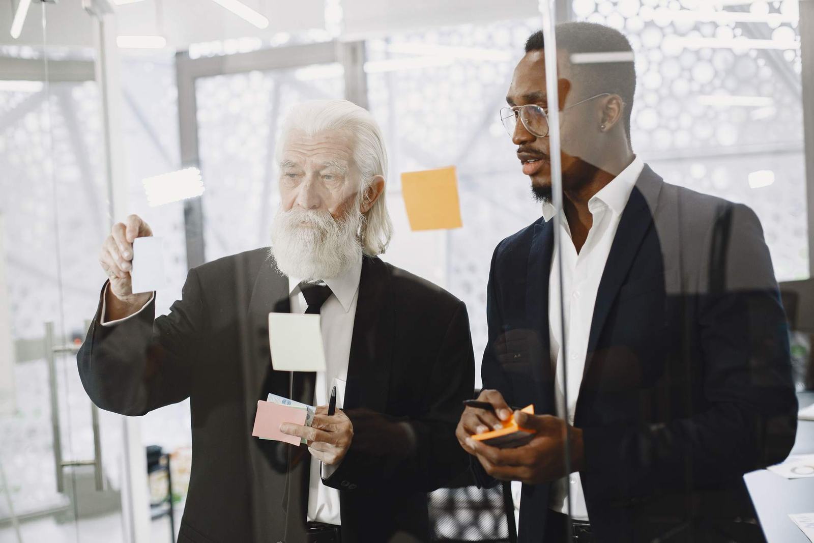 An older consultant and a younger colleague reviewing sticky notes together through a glass wall