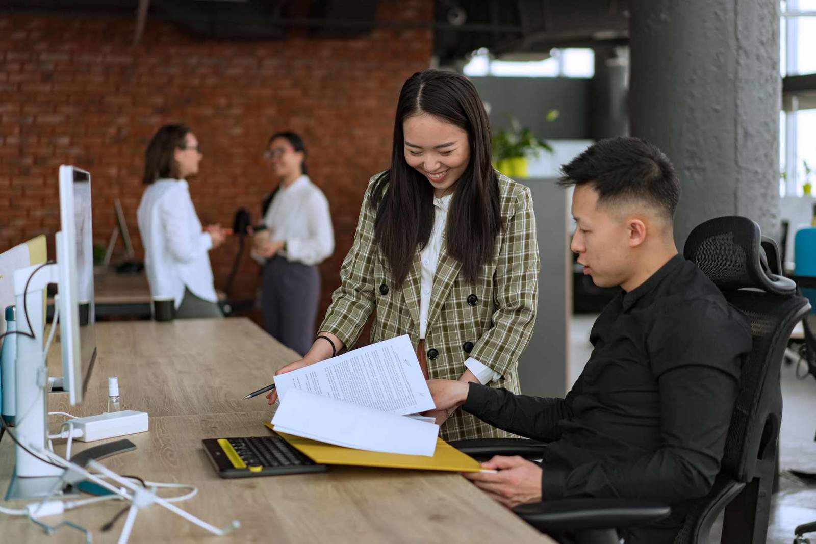A colleague handing over printed documents to a seated coworker at a desk in a bright loft office
