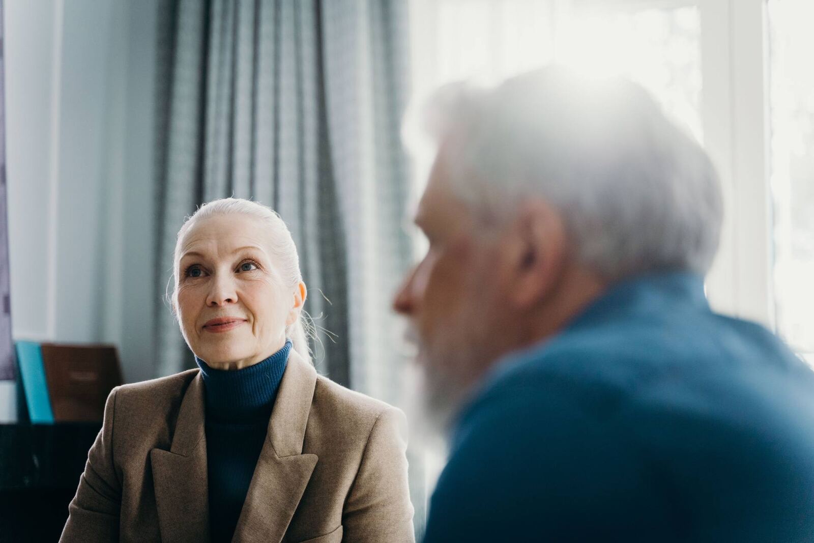 Senior regulatory advisor in a beige blazer and blue turtleneck listening closely to a colleague in soft window light