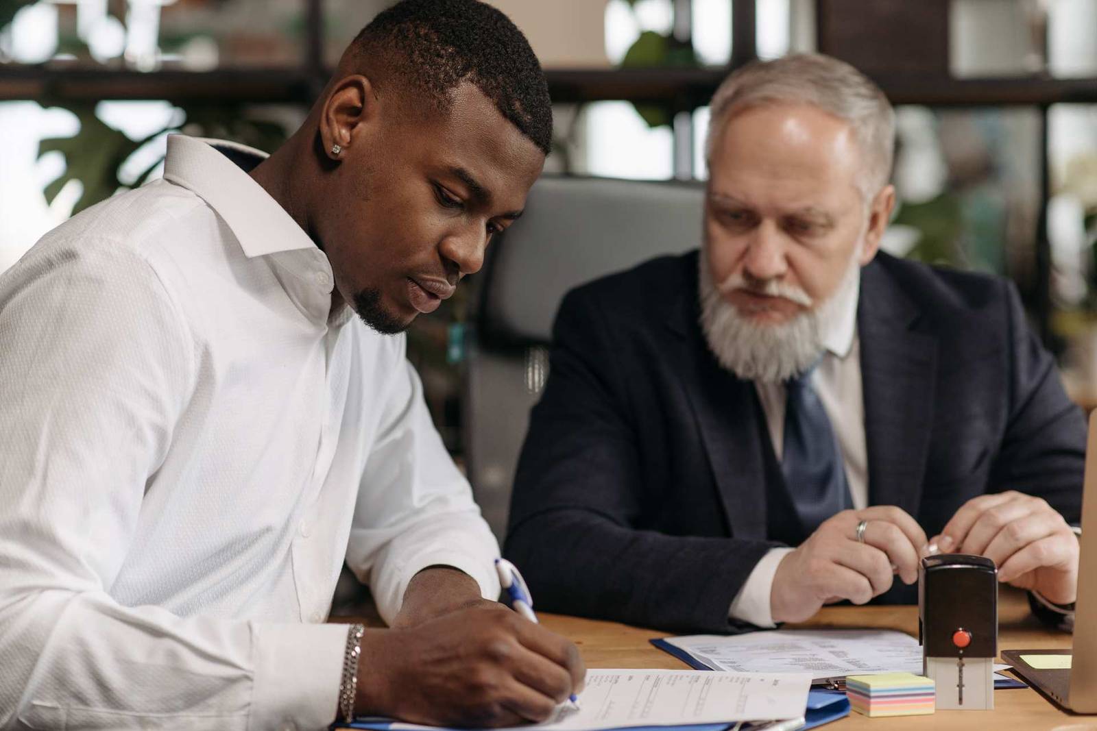 A senior advisor and colleague reviewing a document together at a desk, one hand on a stamp