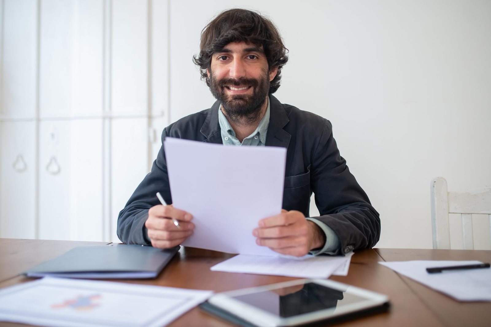 A smiling man in a blazer reviews a paper document at a wooden desk covered with folders, printed charts, and a tablet