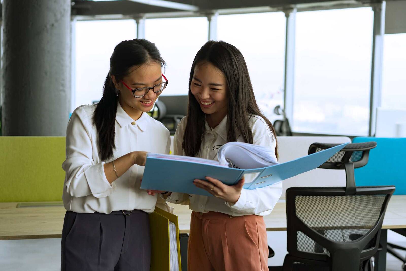 Two colleagues smiling while reviewing an open binder of documents together in a bright, window-lit office
