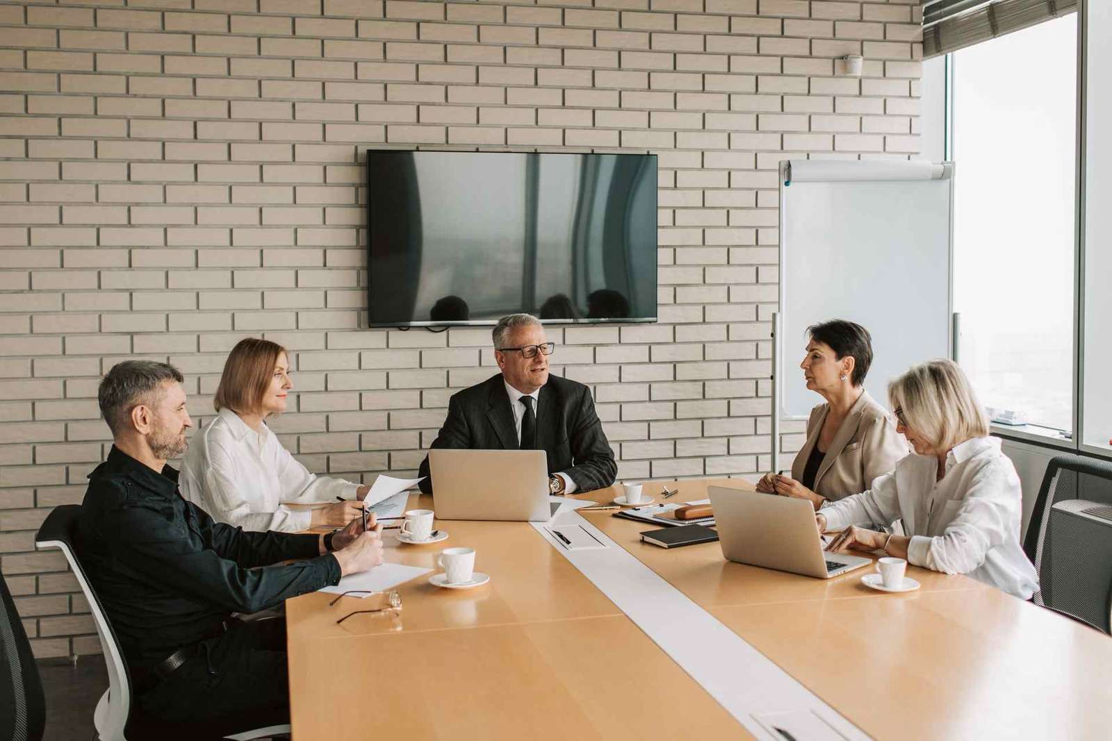 Two colleagues in business attire discussing documents at a wooden conference table with a monitor behind them