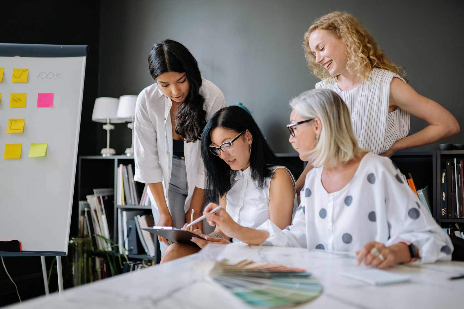 Four colleagues in a bright studio review a clipboard and a fan of color swatches together