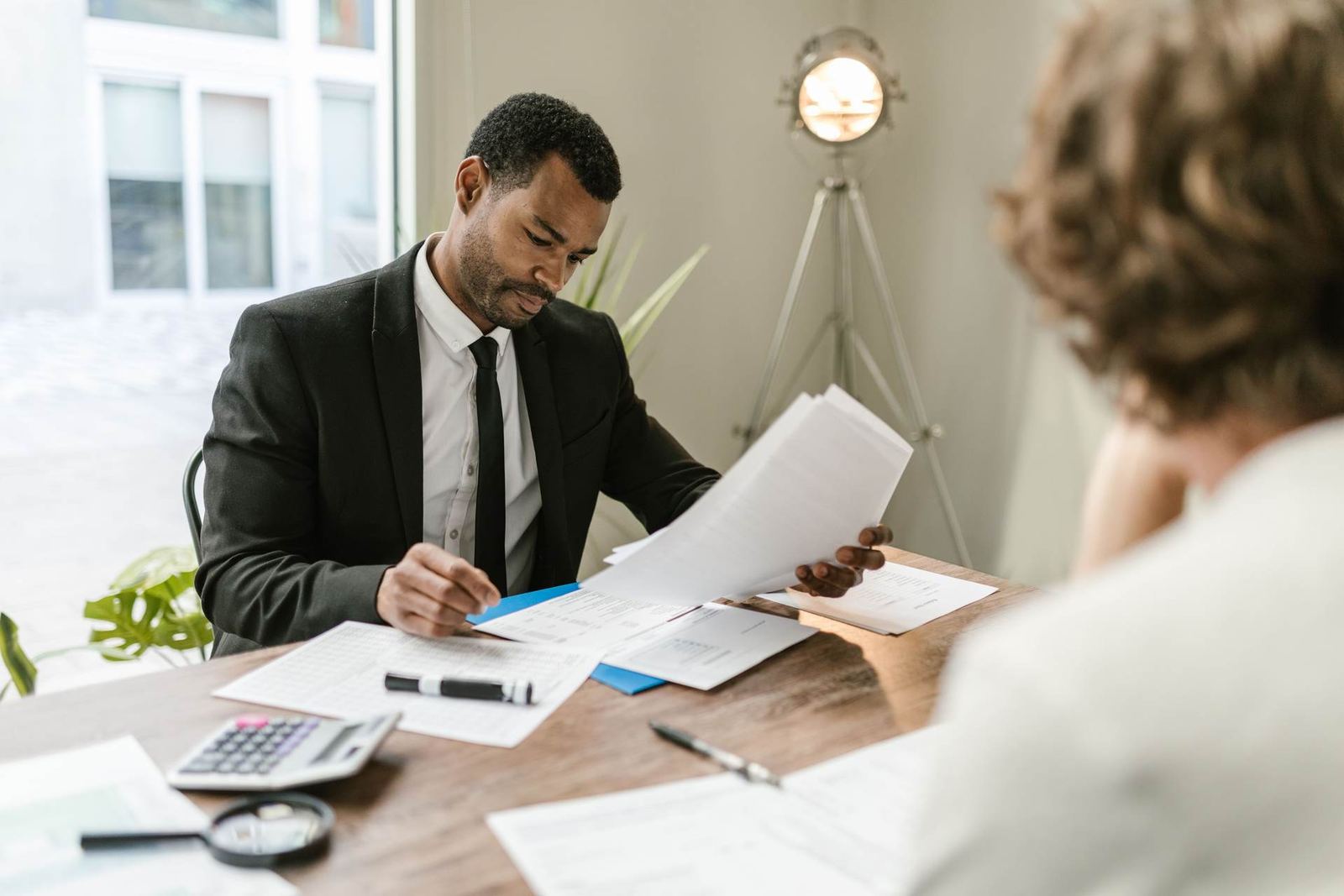 Regulatory professional in a black suit reviewing paperwork across a wooden desk from a colleague