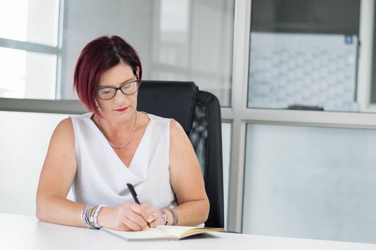 Senior regulatory professional with red hair and glasses writing notes at a white desk in a bright glass-walled office