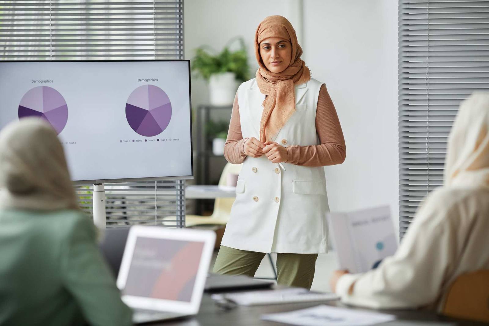 A presenter in a hijab stands before a screen showing demographic pie charts while colleagues look on in a bright meeting room