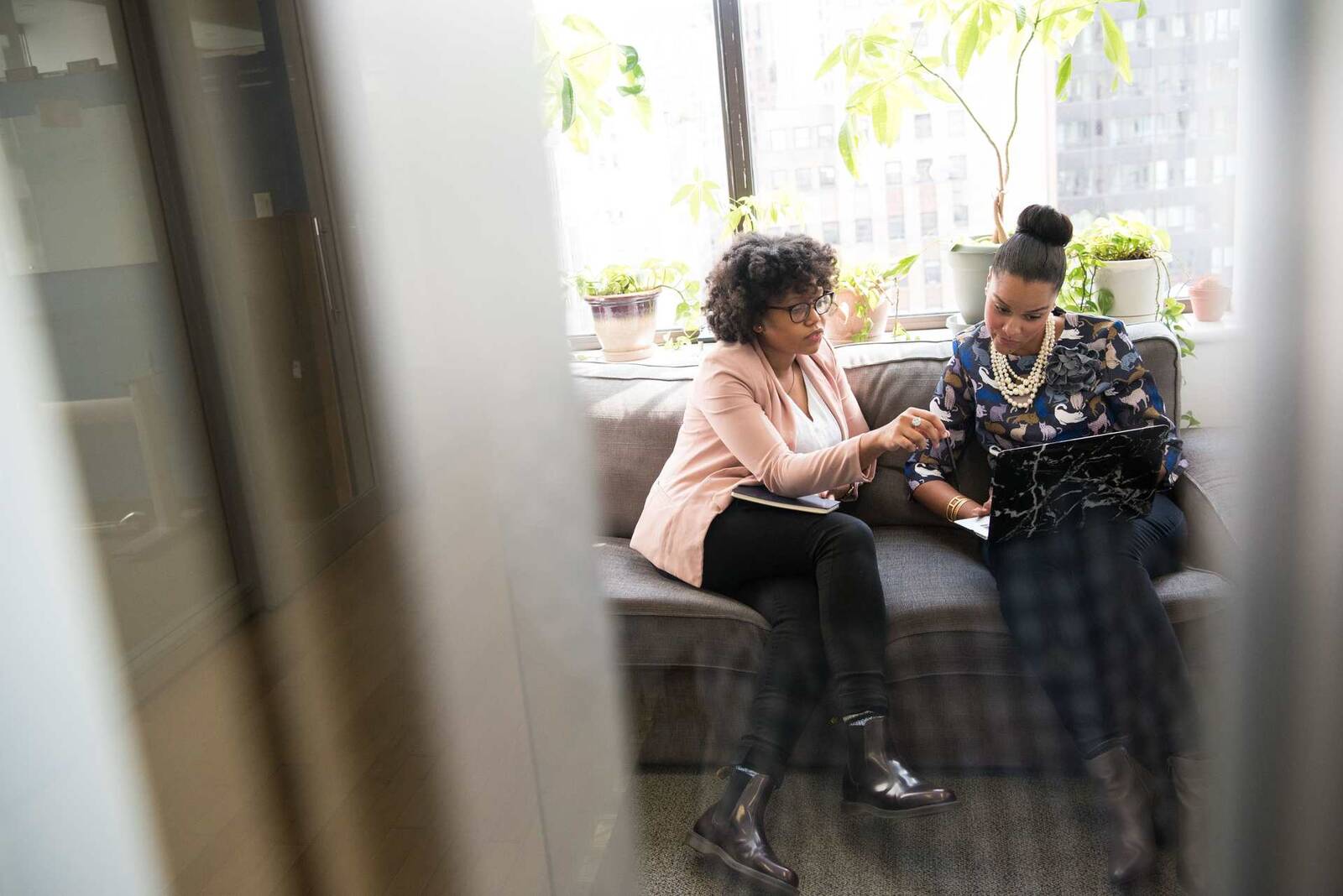 Two professional women review notes together on a sofa in a sunlit high-rise office, with a city skyline and potted plants visible through the window behind them