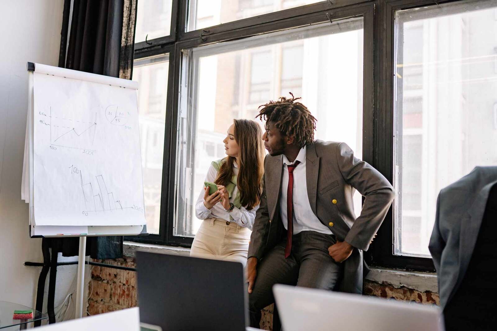Two colleagues reviewing handwritten charts on a flipchart by a window