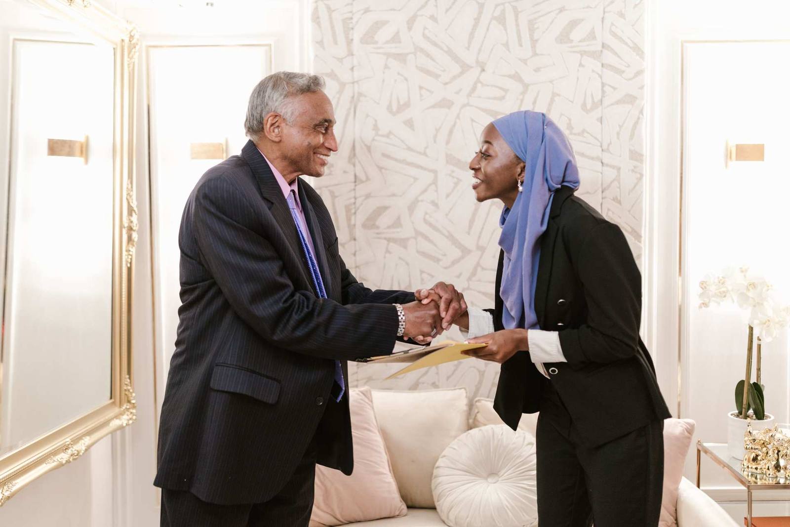 Two regulatory colleagues in business attire shaking hands over a document folder in a bright, elegant office with gold-framed mirrors