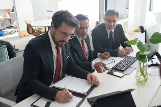 Three colleagues in suits review a marked-up technical document together at a desk with a keyboard and monitor