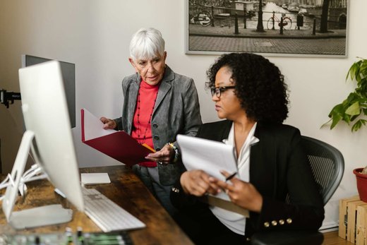 Two colleagues review a red folder of documents together at a desk in a bright office