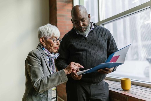 A senior QA leader pointing to a page in an open document folder while a colleague looks on, standing beside a bright office window