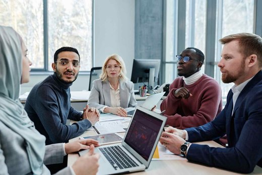 Five colleagues in a bright conference room discussing documents and a laptop together