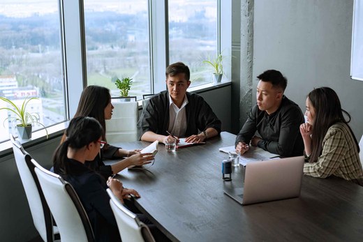Five colleagues gathered around a boardroom table with a laptop, in discussion with a city and park view through floor-to-ceiling windows